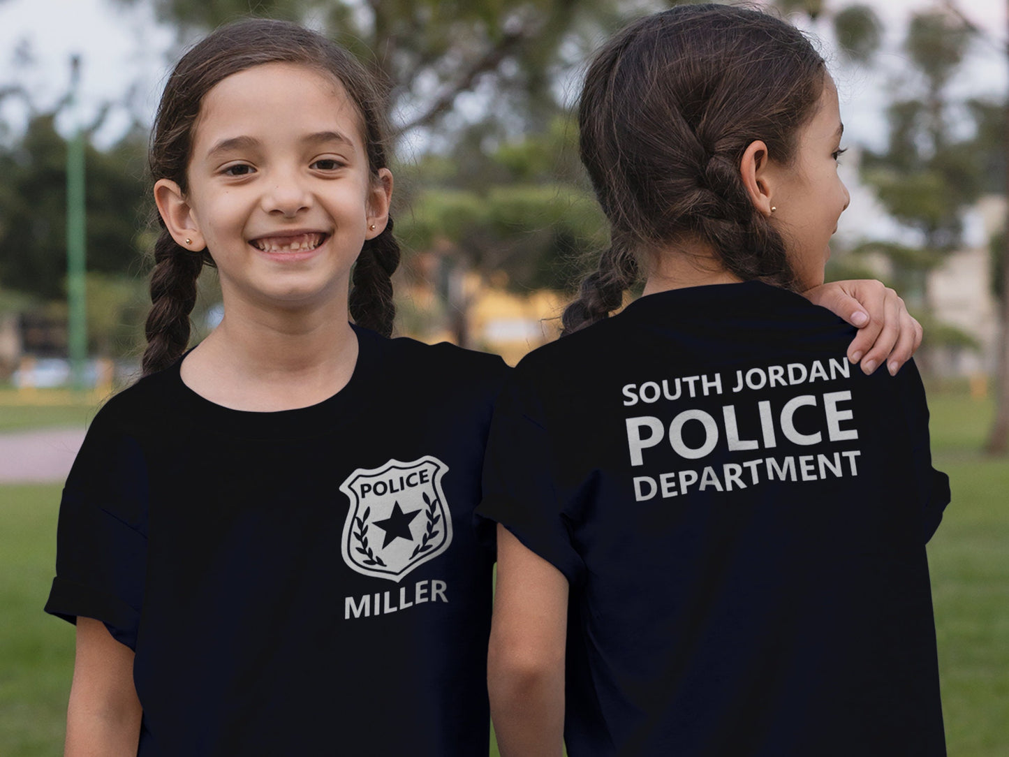 Twin girls wearing black t-shirts with a silver police badge and the name 'MILLER' on the front. The back of the shirt has the text 'SOUTH JORDAN POLICE DEPARTMENT' in silver.