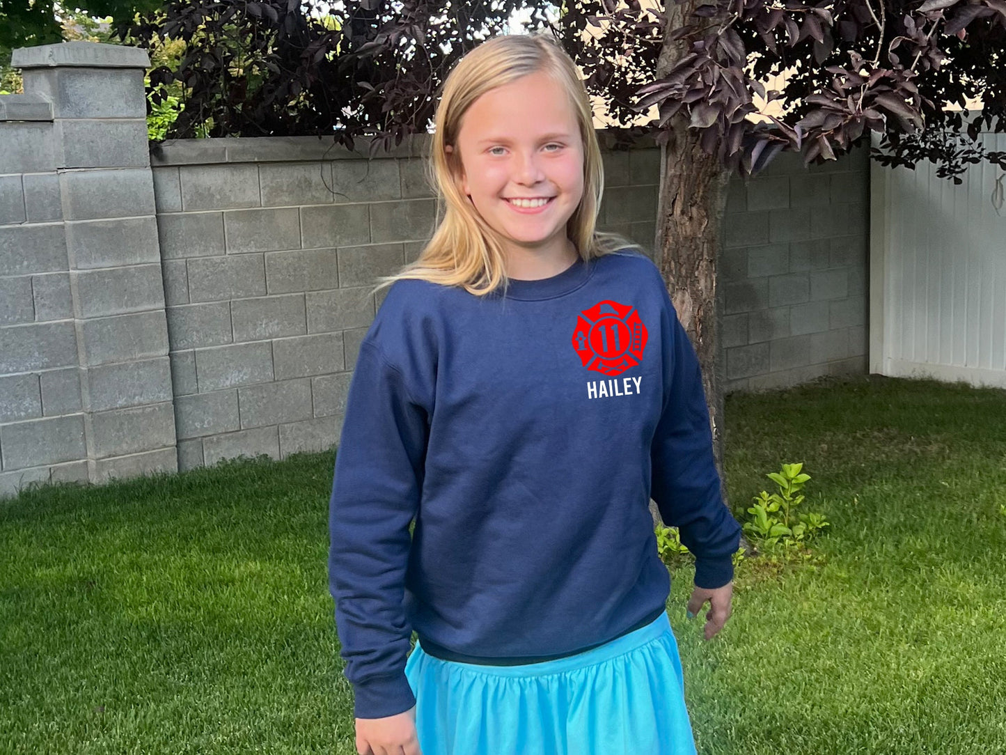 A navy blue sweatshirt with a Maltese Cross graphic and the name 'HAILEY' printed below it. The sweatshirt is worn by a person with long blonde hair.