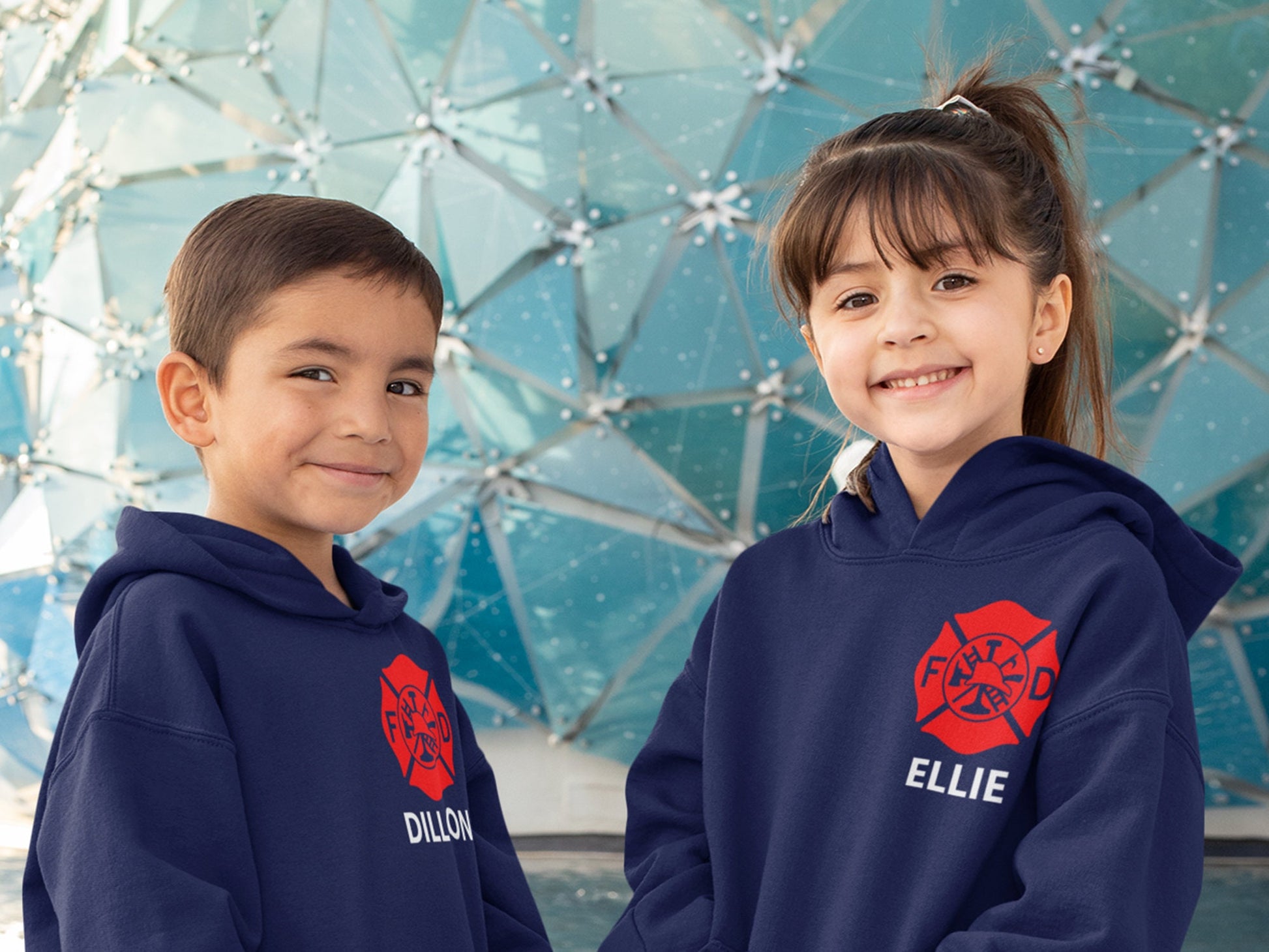 Two children wearing navy blue hoodies with a red firefighter logo and white text. The text on the hoodie on the left says &#39;Dillon&#39; and the text on the hoodie on the right says &#39;Ellie&#39; showing customization option.