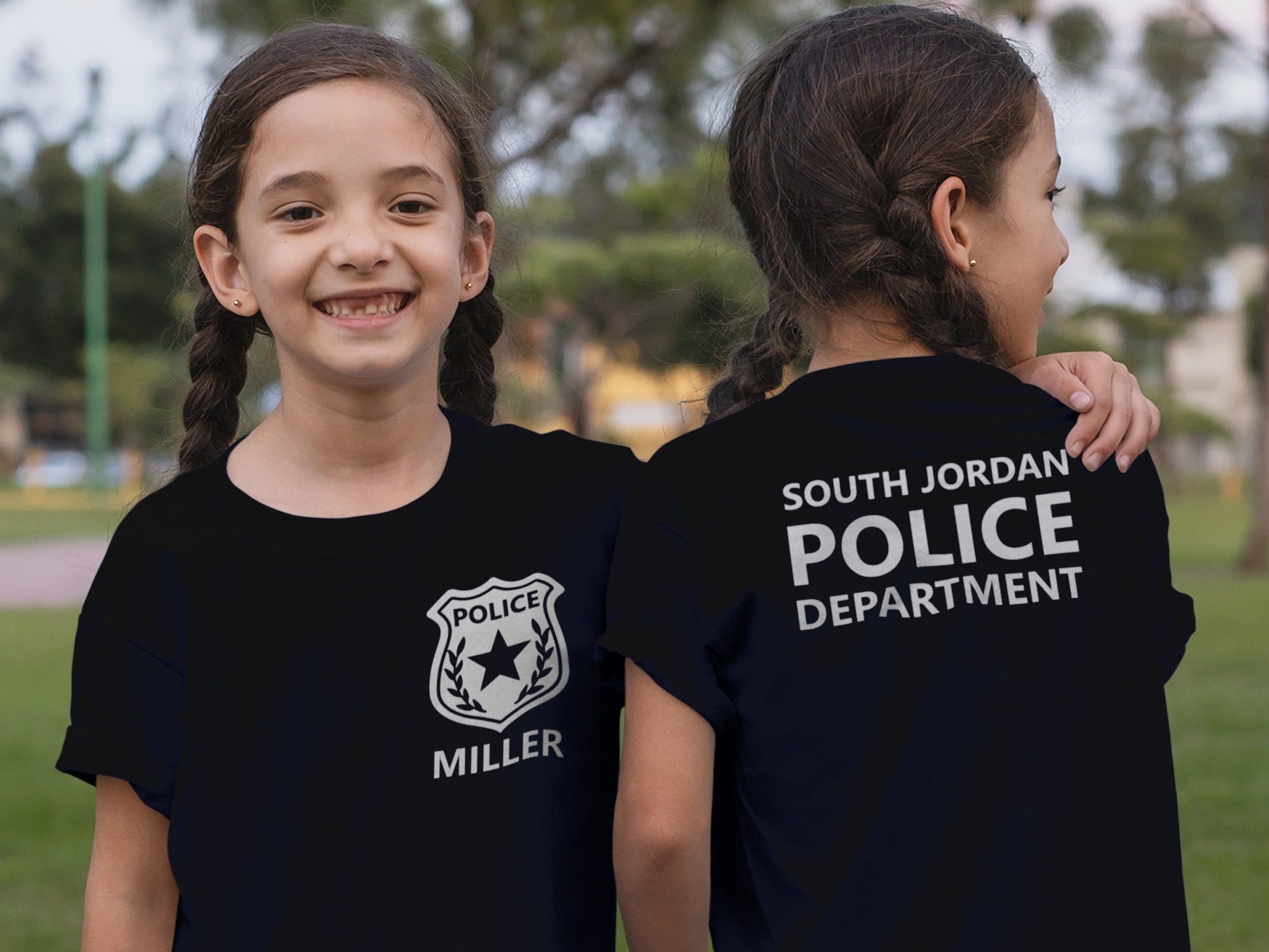 Twin girls wearing black t-shirts with a silver police badge and the name &#39;MILLER&#39; on the front. The back of the shirt has the text &#39;SOUTH JORDAN POLICE DEPARTMENT&#39; in silver.