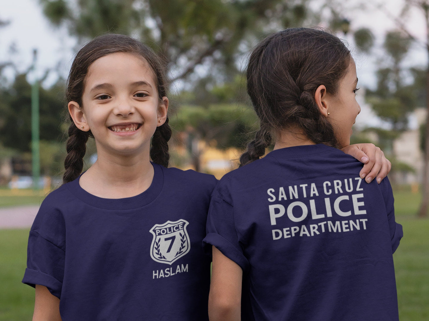 Twin girls wearing navy blue t-shirts. Girl on left showing the front with a silver police badge and number 7 and her name underneath. Left girl showing back of shirt with silver text &quot;santa cruze police department&quot;.