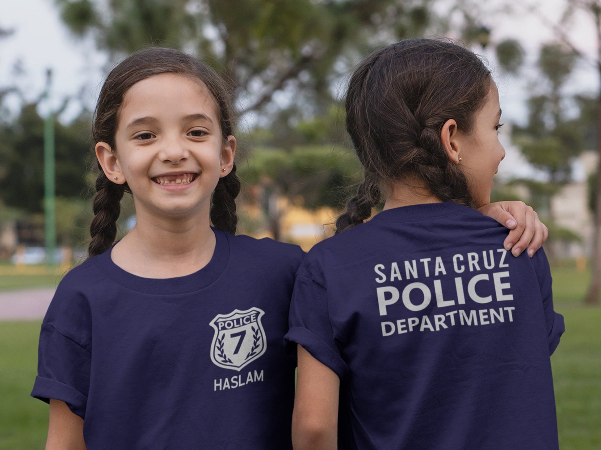Twin girls wearing navy blue t-shirts. Girl on left showing the front with a silver police badge and number 7 and her name underneath. Left girl showing back of shirt with silver text &quot;santa cruze police department&quot;.