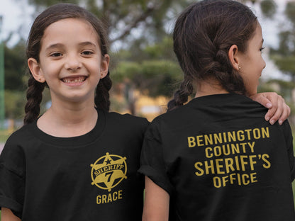Twin girls wearing black t-shirt with a gold sheriff&#39;s star badge on the front and the text &#39;SHERIFF 7 GRACE&#39; underneath. The back of the shirt has the text &#39;BENNINGTON COUNTY SHERIFF&#39;S OFFICE&#39; in gold lettering.