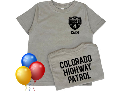 A gray toddler t-shirt with a black state trooper patch on the front that says &#39;State Trooper&#39; with a star on either side of the number &#39;4&#39; and the name &#39;Cash&#39; below. The back of the shirt says &#39;Colorado Highway Patrol&#39; in black text. Colorful balloons indicate a birthday theme.
