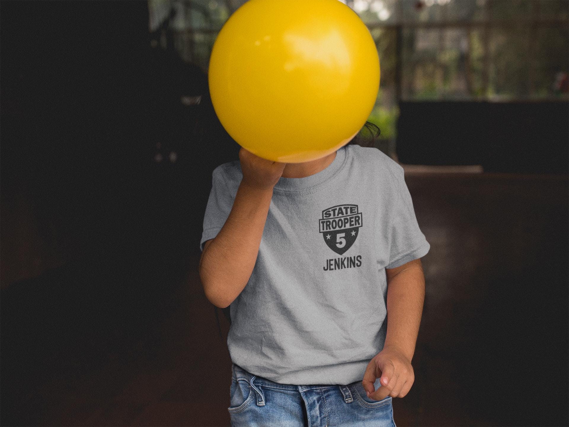 Toddler covering face with a balloon wearing a gray t-shirt with a black badge that says &#39;STATE TROOPER&#39; and the number &#39;5&#39; with stars on each side. The name &#39;JENKINS&#39; is underneath in black text.
