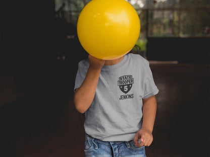 Toddler covering face with a balloon wearing a gray t-shirt with a black badge that says &#39;STATE TROOPER&#39; and the number &#39;5&#39; with stars on each side. The name &#39;JENKINS&#39; is underneath in black text.