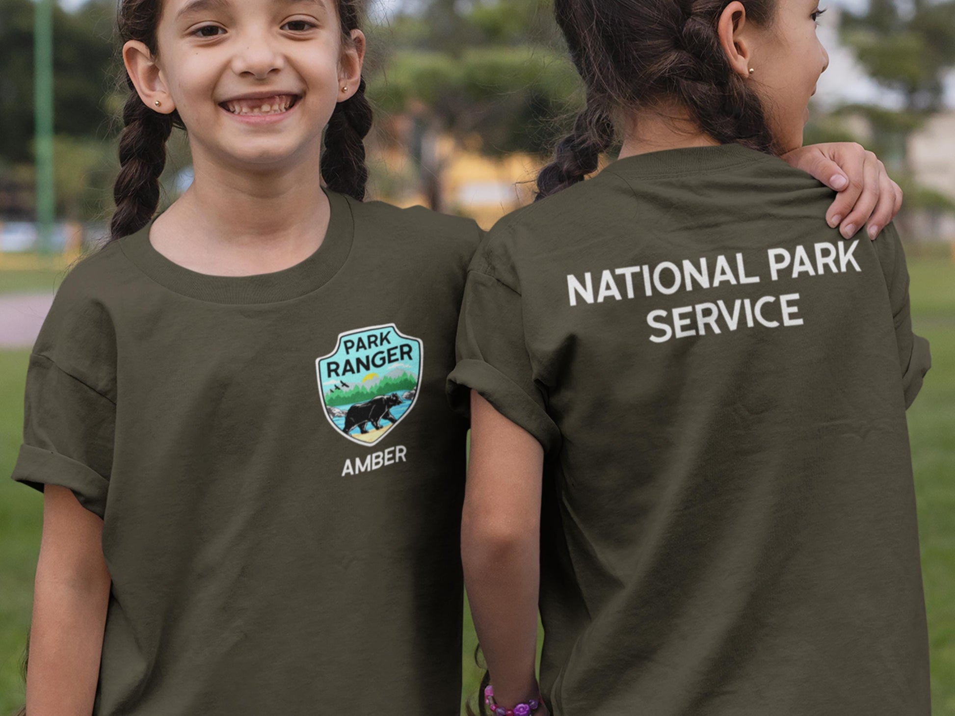 Two children wearing dark green t-shirts. The t-shirt on the left has a green, blue, and yellow patch with a bear and the text &#39;Park Ranger&#39; and &#39;Amber&#39;. The t-shirt on the right has the text &#39;National Park Service&#39; on the back.