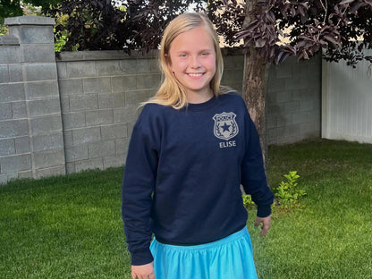 Girl wearing a navy blue sweatshirt with a silver police badge emblem on the left chest and the name &#39;HAILEY&#39; in silver text.