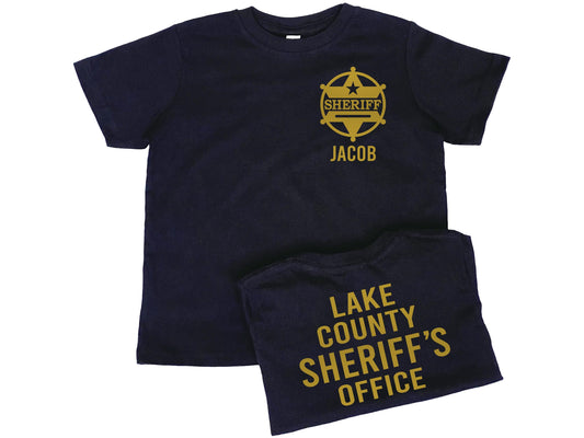 Black toddler t-shirt sporting a gold sheriff badge with 6 pointed star and a ring around it. The badge also has a 5 pointed star above the word Sheriff and the name &quot;Jacob&quot; underneath. The back of the shirt has gold lettering &quot;Lake County Sheriff&#39;s Office&quot; as a place holder for personalized Text.