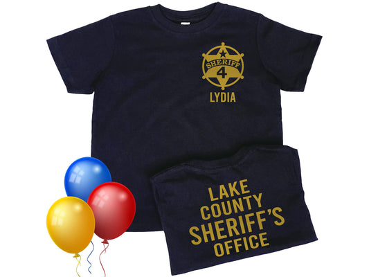Toddler black t-shirt with told star sheriff badge on the left chest. Badge contains the number 4 in the center and the name &#39;Lydia&#39; underneath as placeholders for child&#39;s name and age. The back of the shirt says &#39;Lake County Sheriff&#39;s Office&#39; in gold lettering.